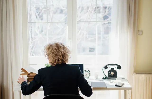 Person seated with an open book at a desk beside a laptop, telephone and sunlit window