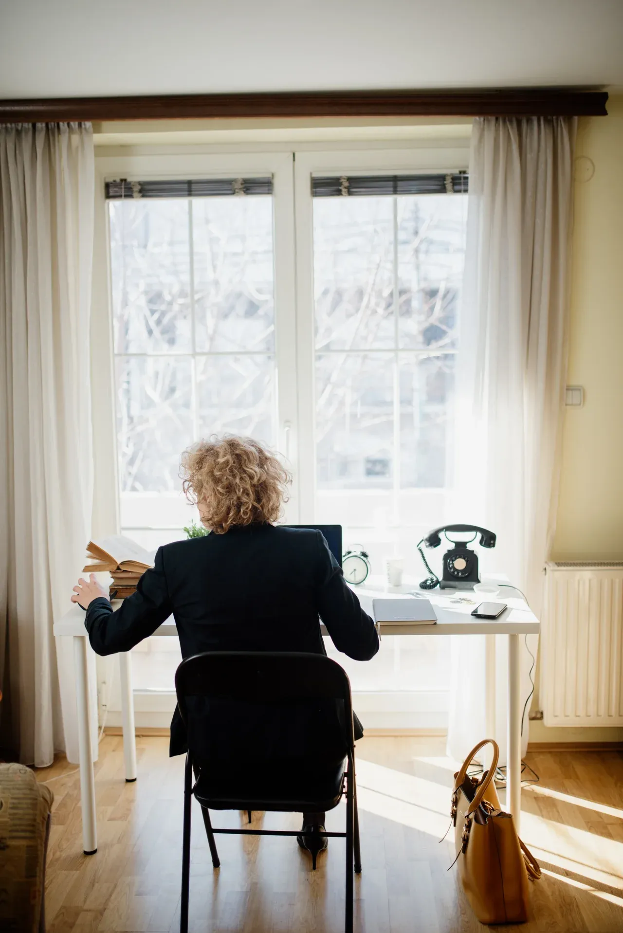 Person seated with an open book at a desk beside a laptop, telephone and sunlit window