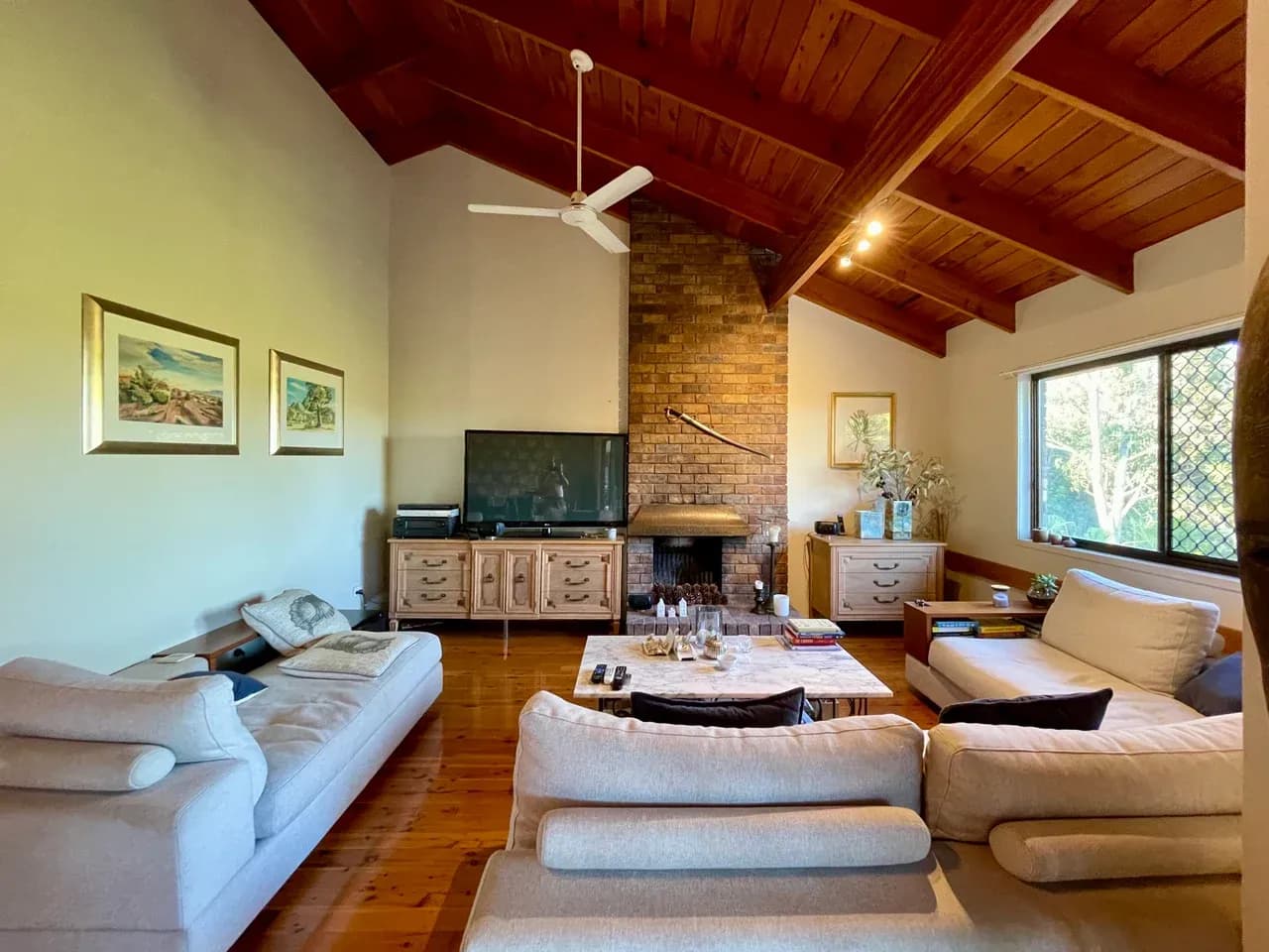 Furnished sitting room in Corinda, Queensland, Australia with sofas, a brick fireplace, exposed ceiling beams and a window