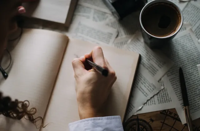 Hand writing in an open notebook beside scattered printed pages, a camera and a drink