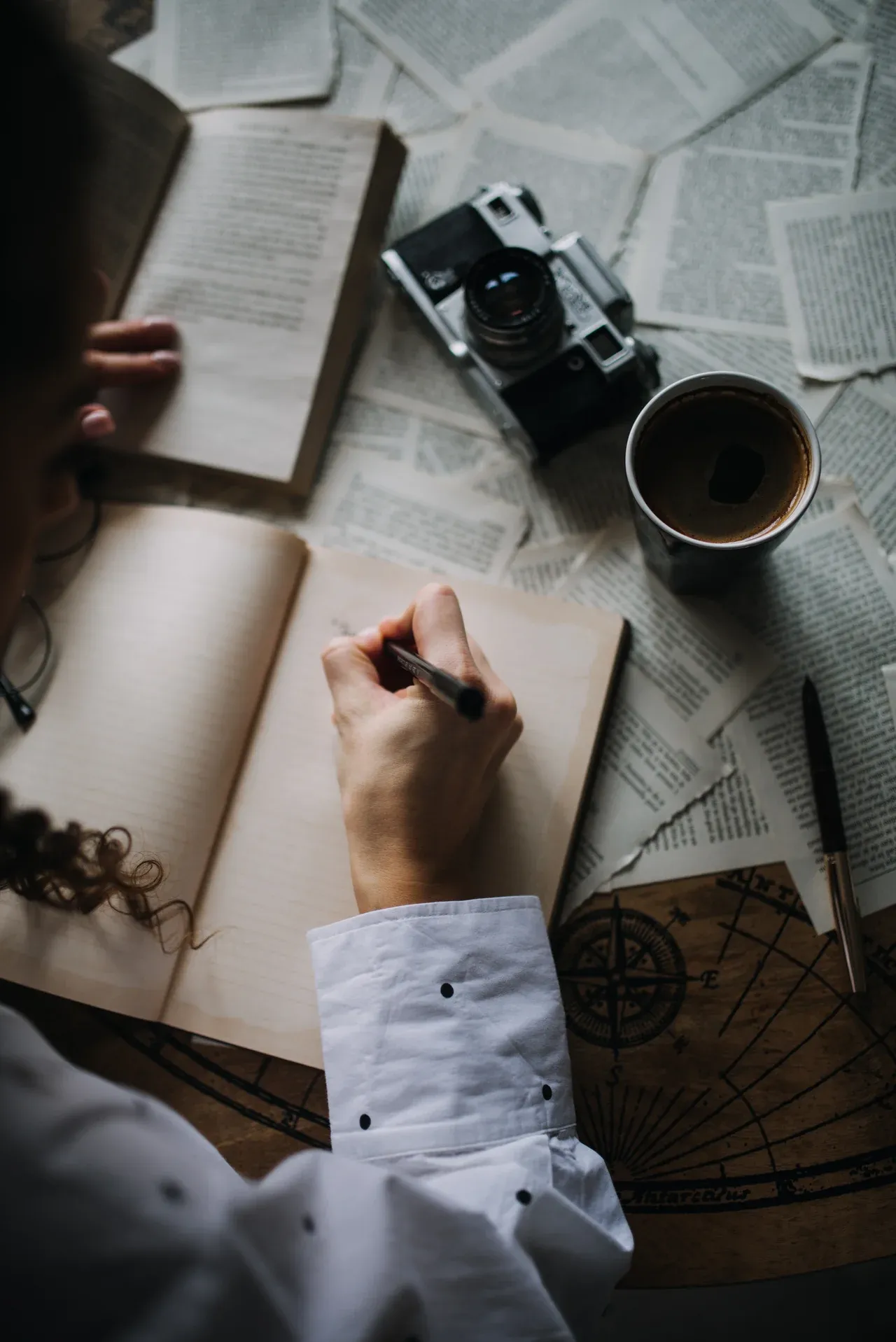 Hand writing in an open notebook beside scattered printed pages, a camera and a drink