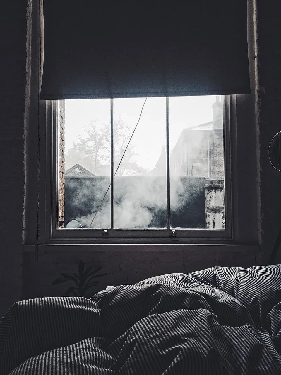 Bedroom window with a partly lowered blind and nearby rooftops visible beyond the glass