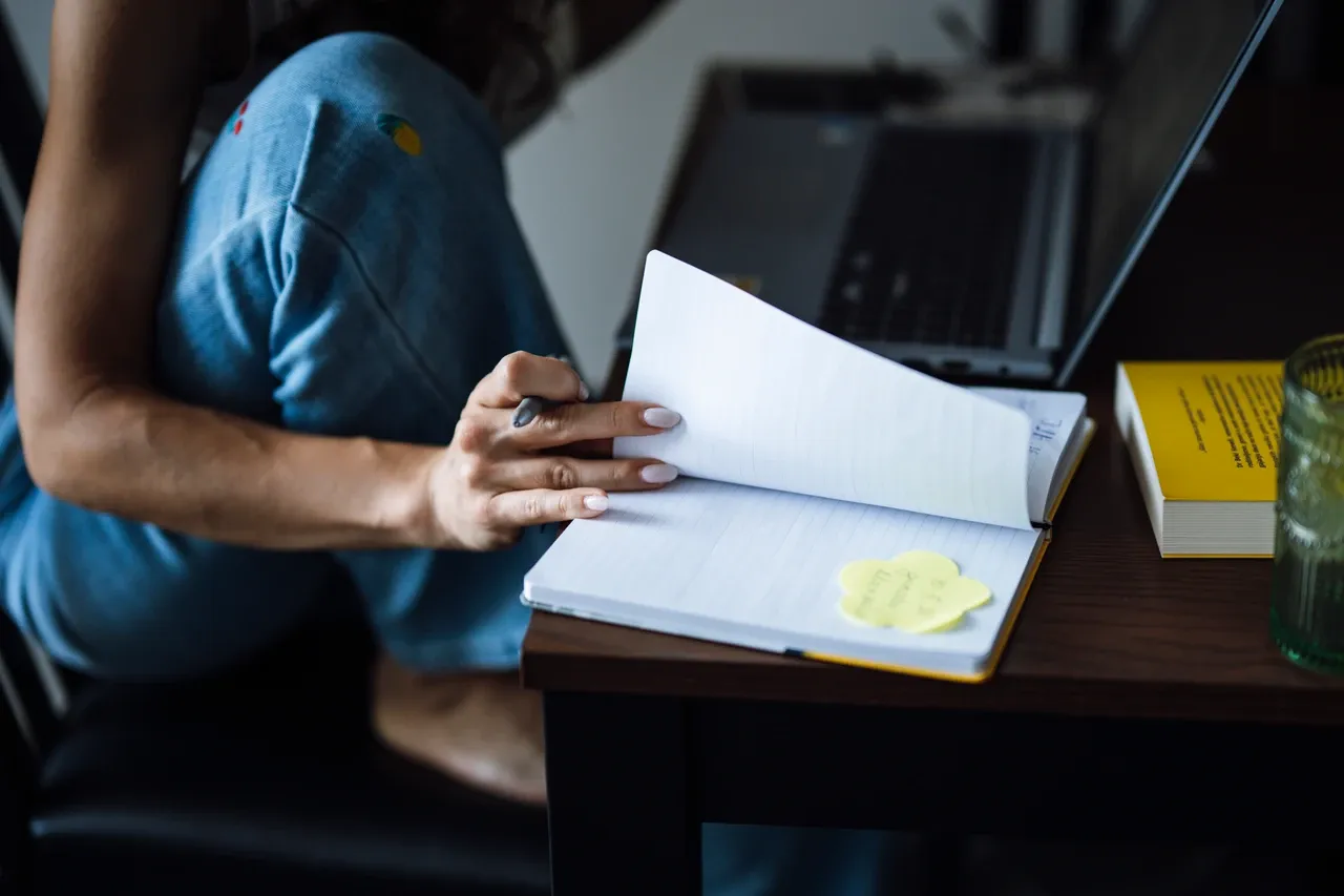 Person holding a pen and turning a notebook page beside a laptop on a wooden table, photographed in June 2026