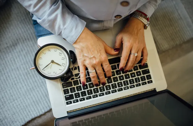 Hands typing on a laptop with an analogue alarm clock resting beside the keyboard, photographed in October 2021