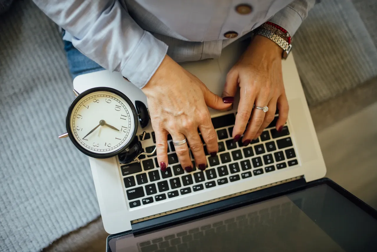 Hands typing on a laptop with an analogue alarm clock resting beside the keyboard, photographed in October 2021