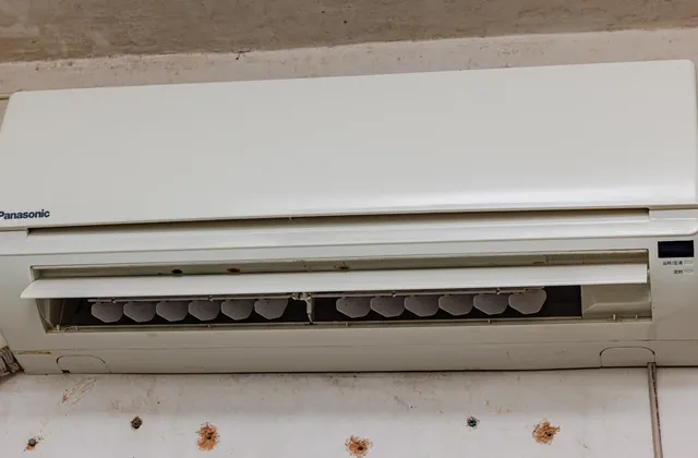 White wall-mounted air-conditioning unit with an open outlet and louvres above a marked wall, with a pipe visible at the left