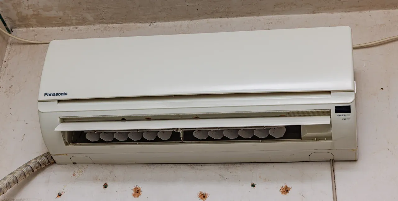 White wall-mounted air-conditioning unit with an open outlet and louvres above a marked wall, with a pipe visible at the left