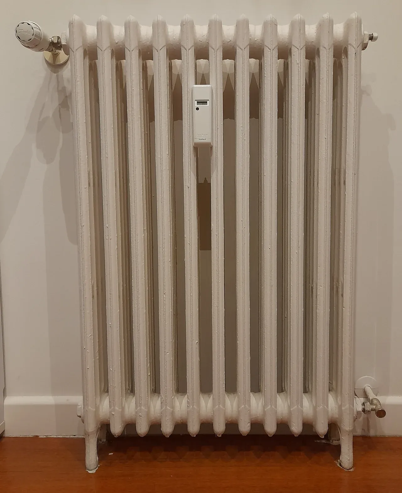 Cream-coloured radiator with a control knob, a small attached display device and visible pipes above a wood-effect floor