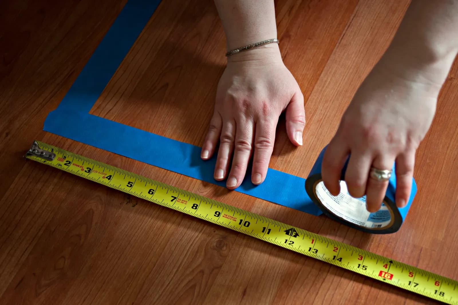 Hands marking a wooden floor area using blue tape and a yellow measuring tape in an illustrative 2014 photograph