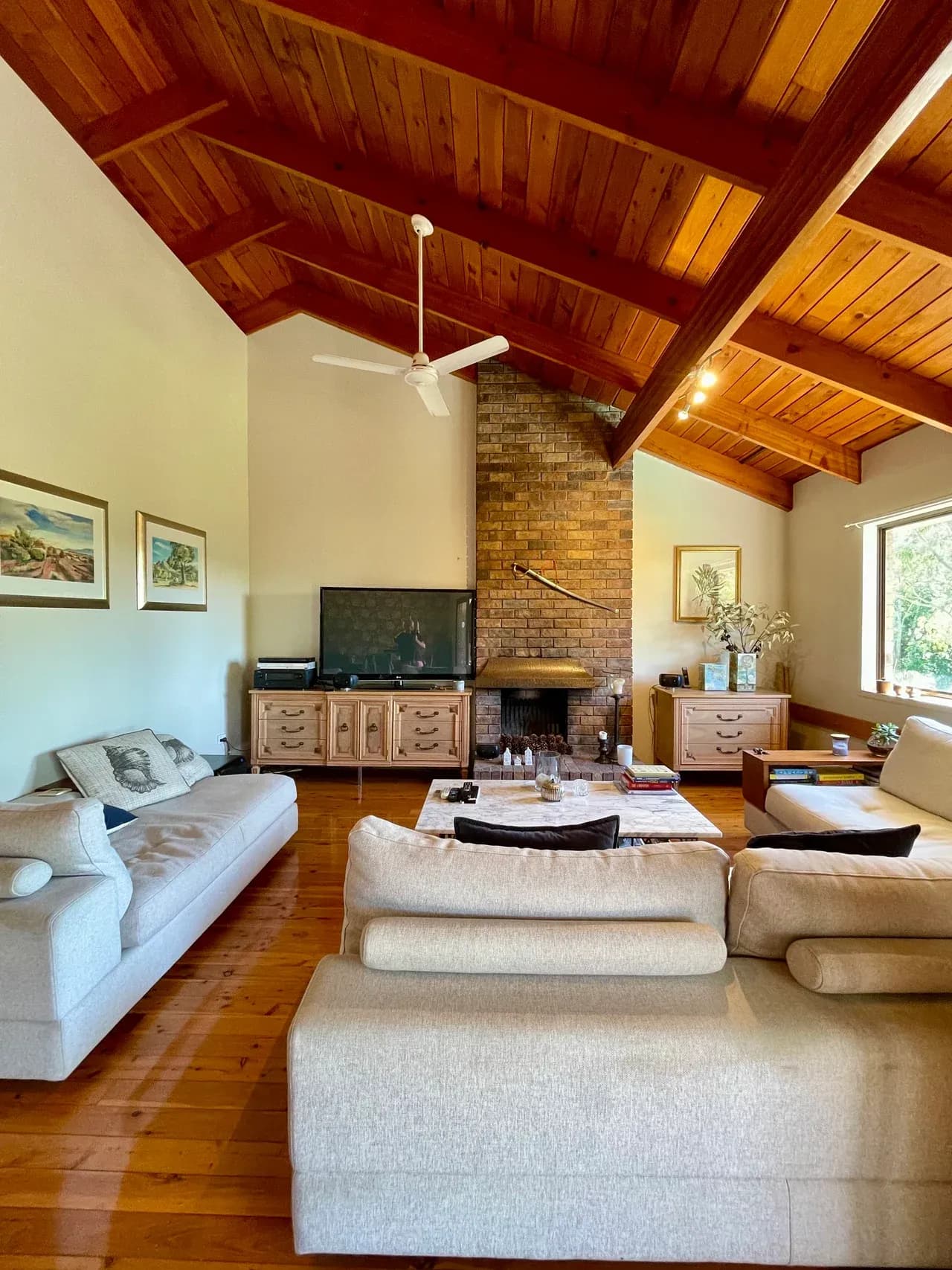 Furnished sitting room with a high timber-lined ceiling, brick fireplace, sofas and window in Corinda, Queensland, Australia