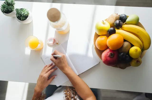 Hands writing in an open notebook beside a fruit platter and juice on a white table