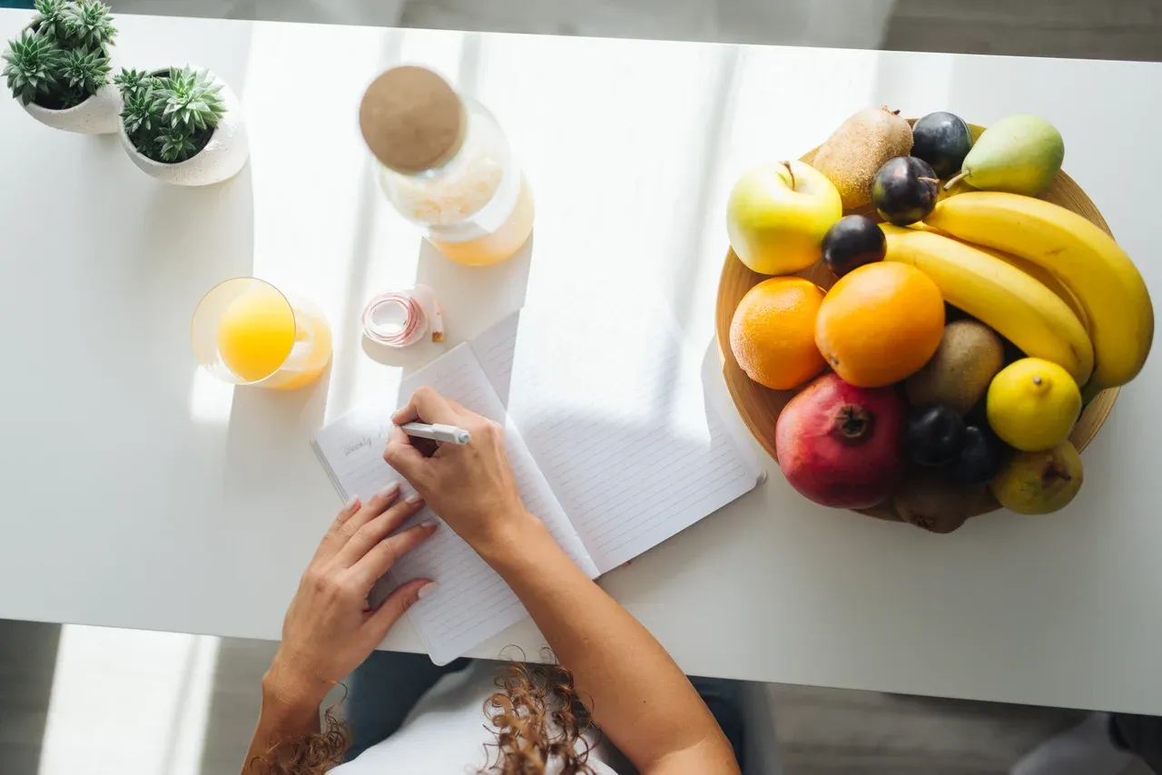 Hands writing in an open notebook beside a fruit platter and juice on a white table