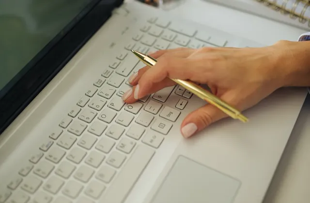 Hand holding a gold-coloured pen over a laptop keyboard, with notebooks and a phone on the desk