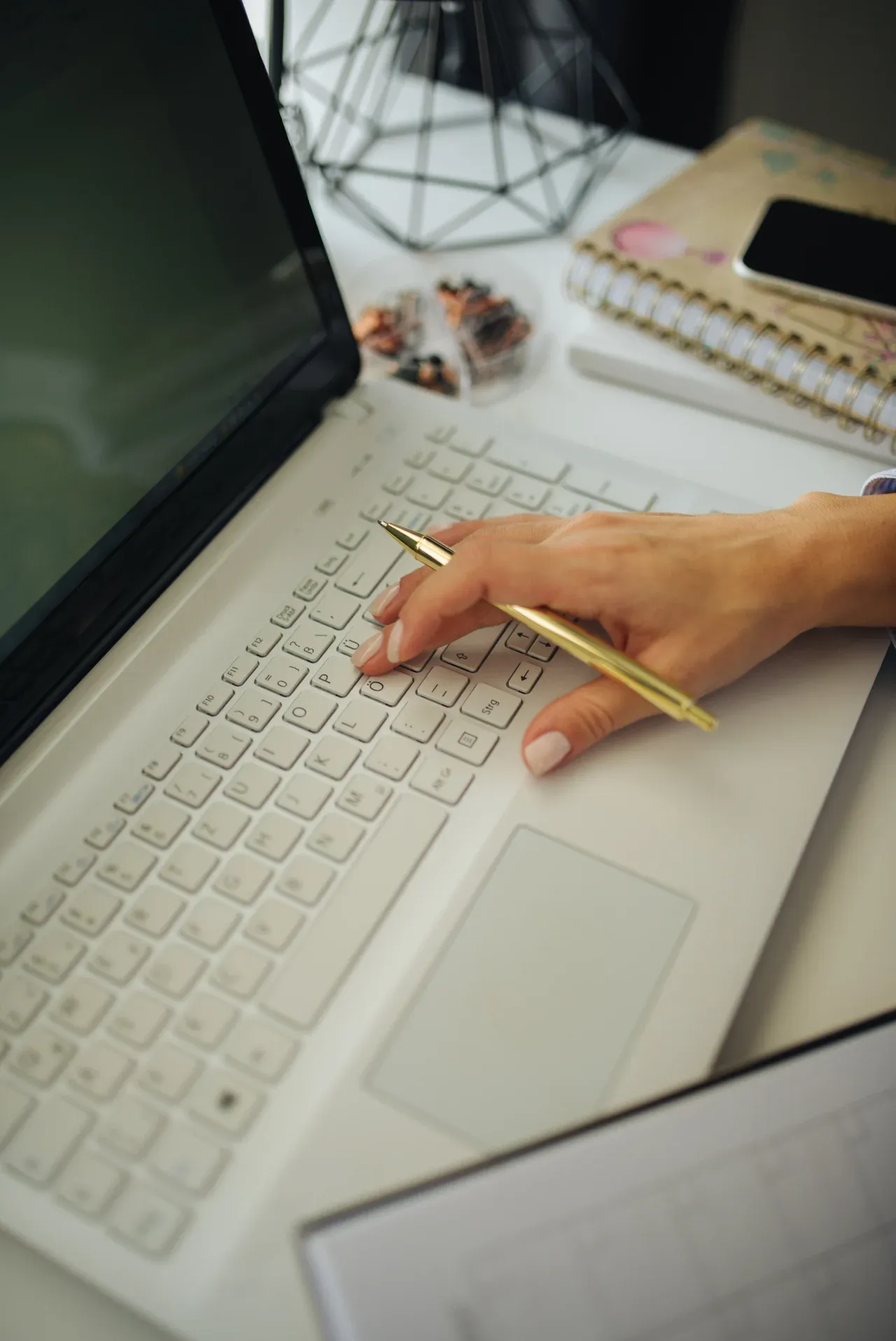 Hand holding a gold-coloured pen over a laptop keyboard, with notebooks and a phone on the desk