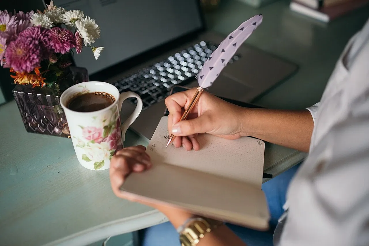 Hand writing in a notebook with a feather pen beside coffee and flowers
