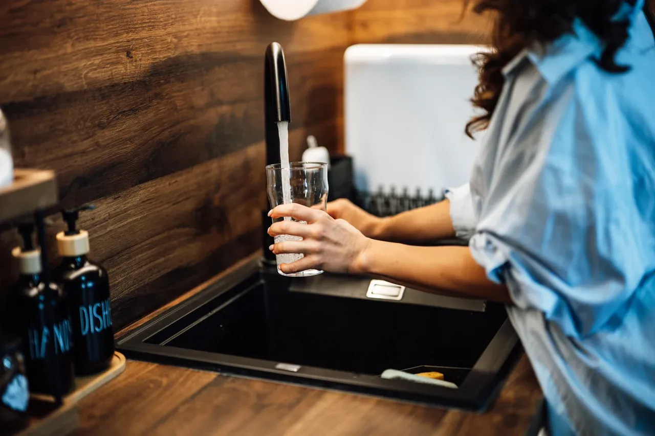 Person holding a glass under flowing water from a kitchen tap above a dark sink