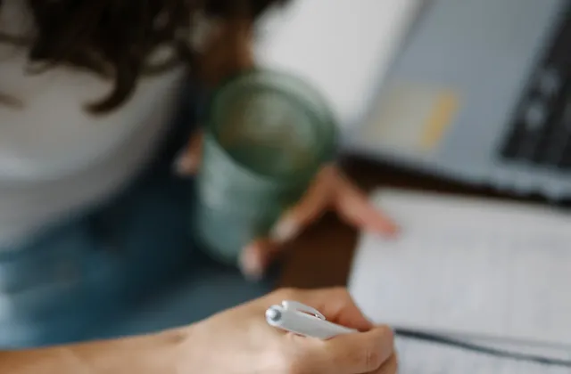 Person writing in an open notebook while holding a drink beside a laptop