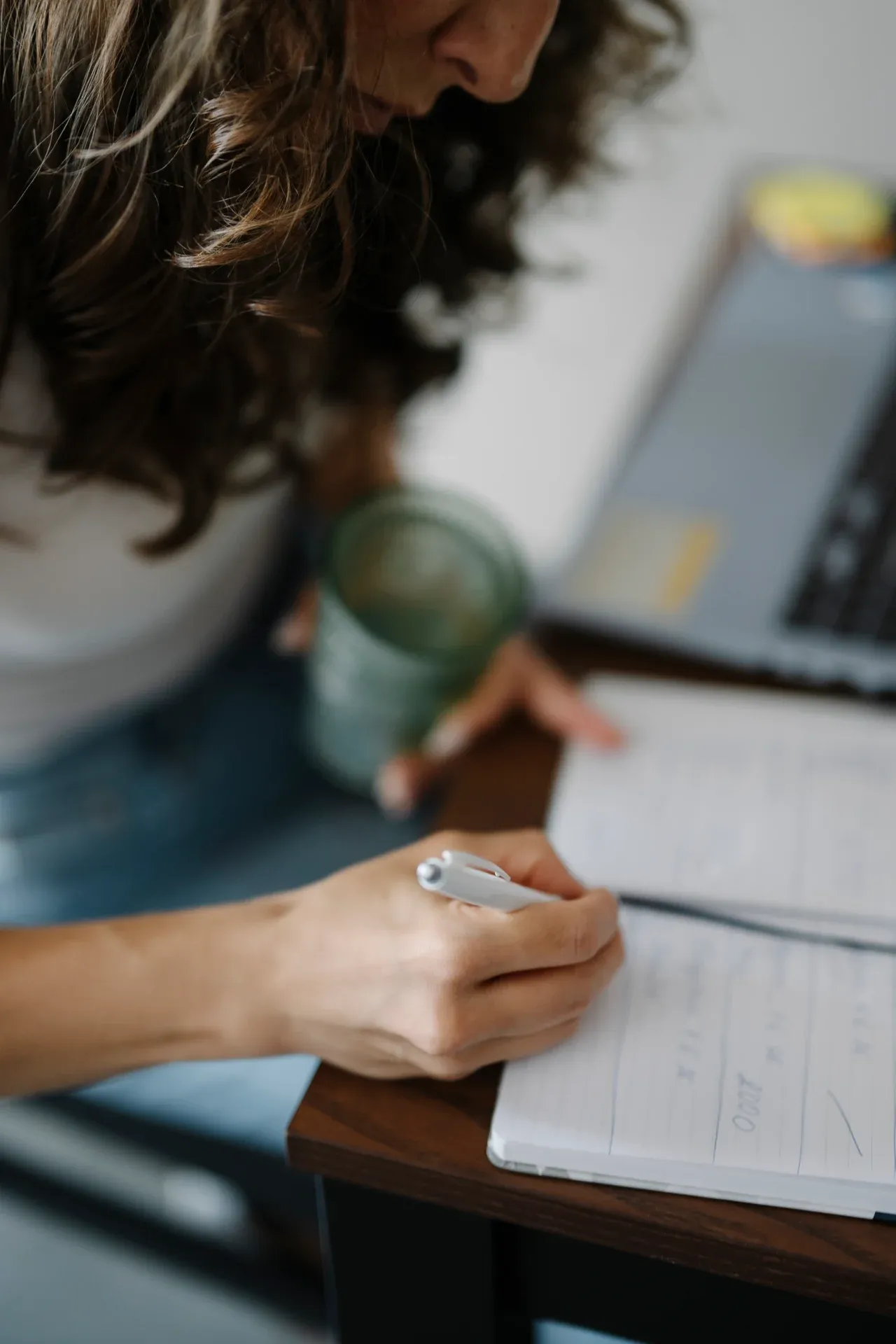 Person writing in an open notebook while holding a drink beside a laptop