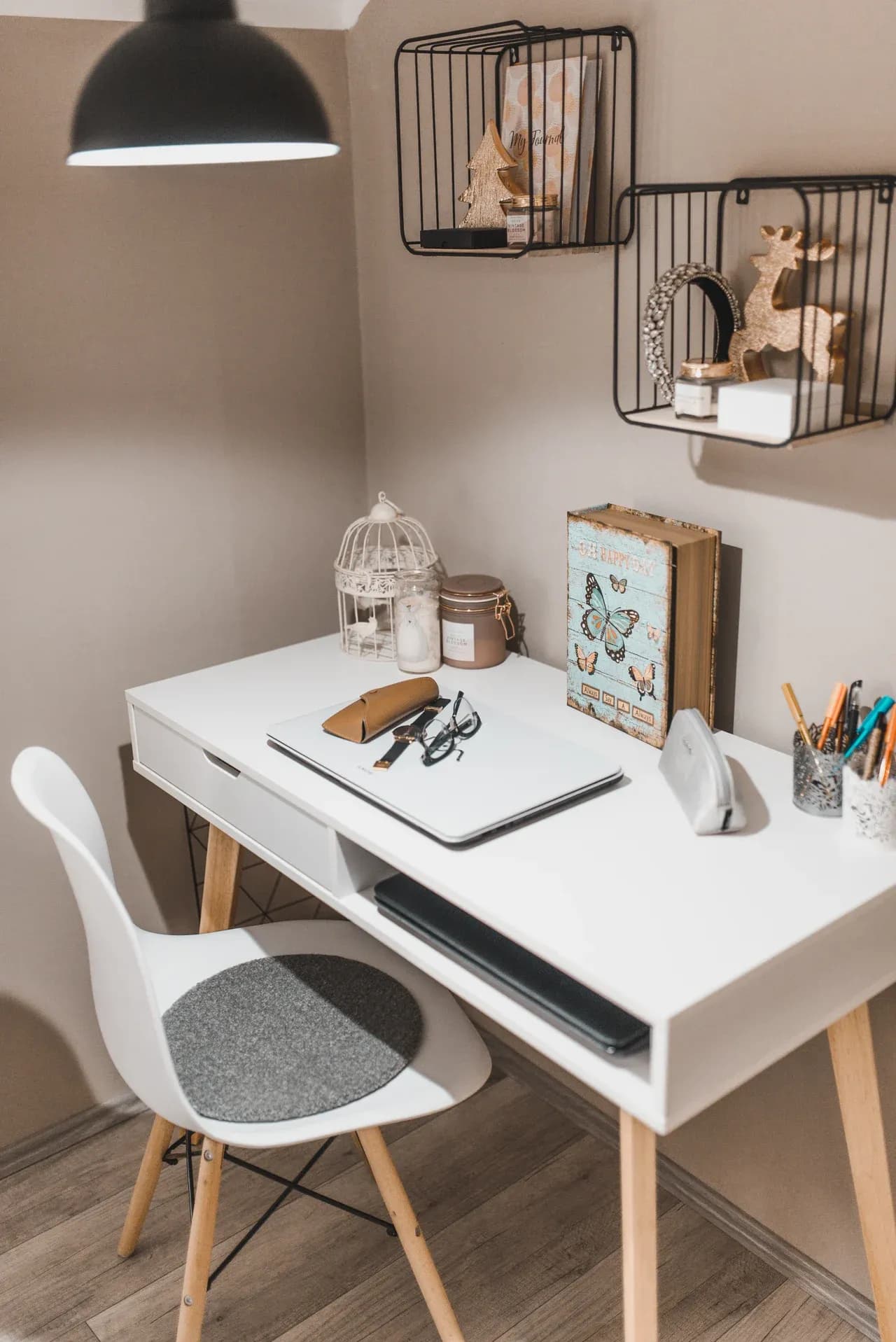 White desk with a closed laptop, glasses, keys and pen containers beneath wall shelves and a hanging lamp