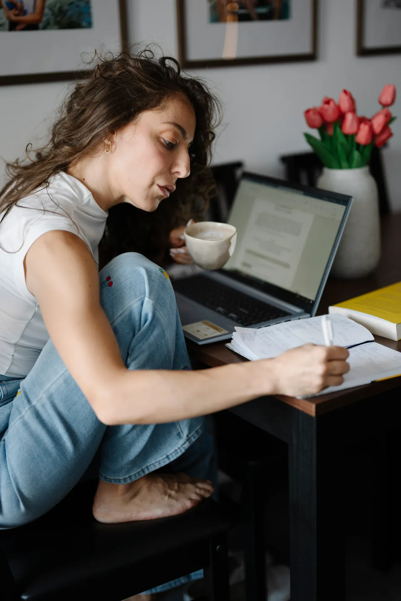 Woman holding a cup and writing in an open notebook beside a laptop and flowers