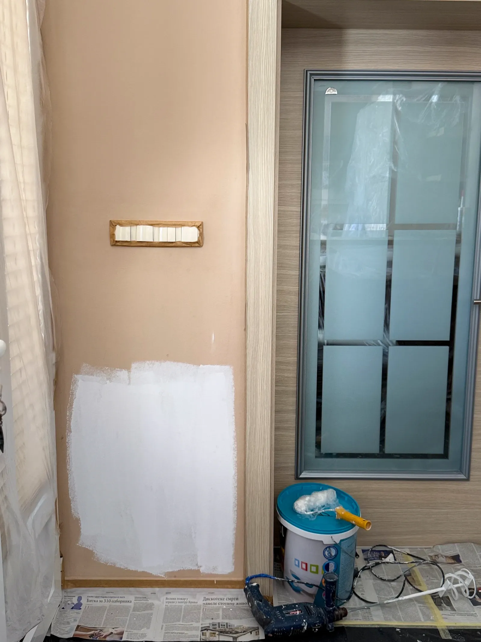 Partly painted interior wall beside a door, with a paint container and tools, May 2026