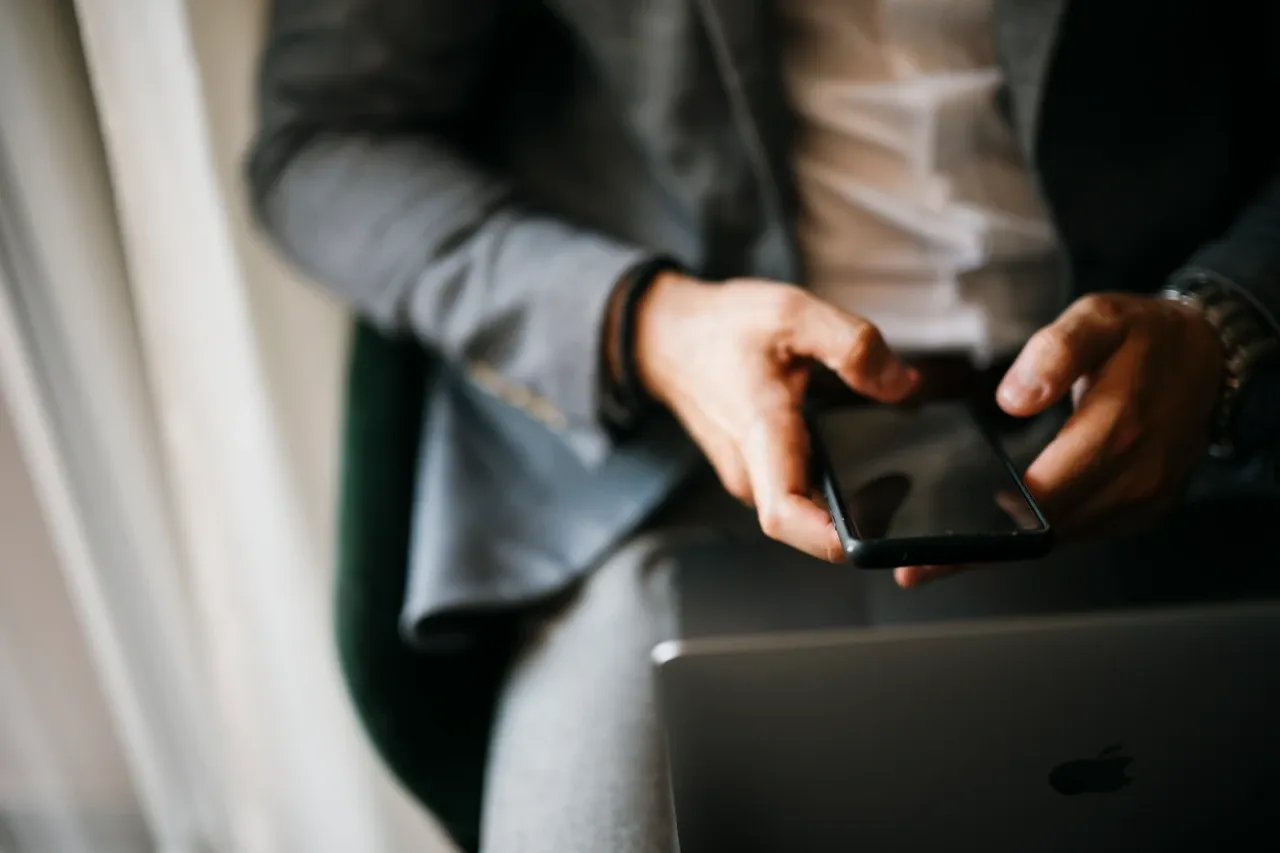 Hands holding a smartphone beside an open laptop, photographed in June 2026