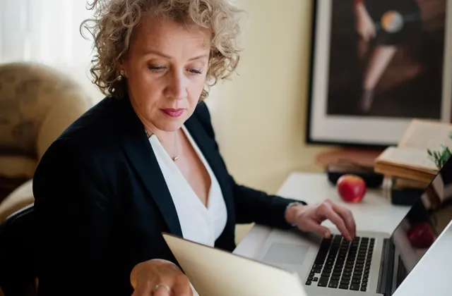 Woman looking at an open notebook with one hand on a laptop beside a sunlit window