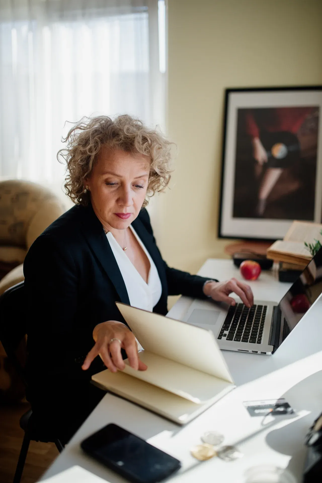 Woman looking at an open notebook with one hand on a laptop beside a sunlit window