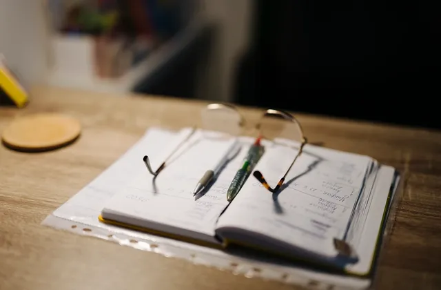 Open notebook with two pens and reading glasses on a wooden desk, photographed in June 2026