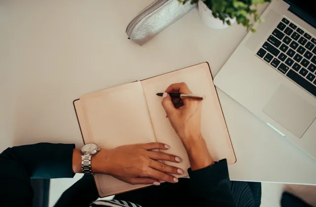 Hands holding an open lined notebook and pen beside a laptop keyboard on a white desk