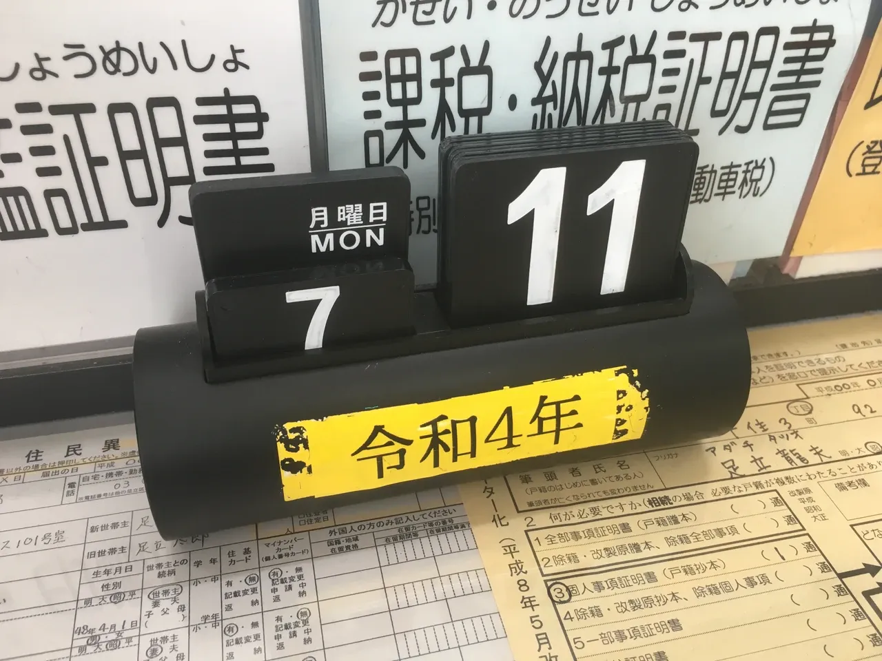 Black desktop calendar displaying Monday, July 11, beside Japanese-language signs and printed sheets
