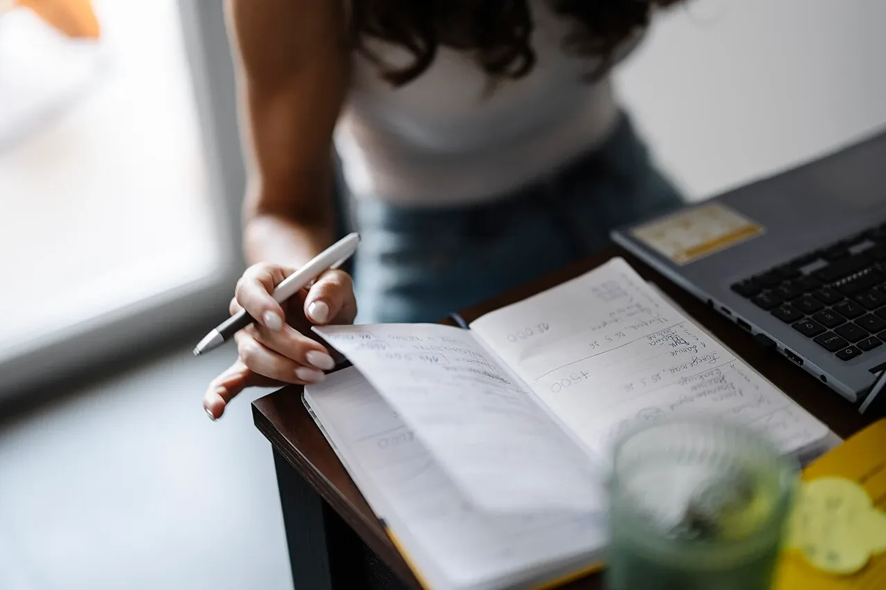 Hand holding a pen over a notebook beside a laptop and a glass of water, photographed in June 2026