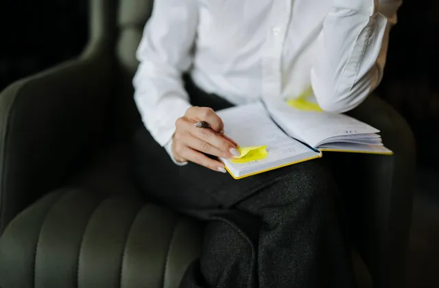 Person writing in an open notebook with sticky notes while seated in an armchair, photographed in June 2026