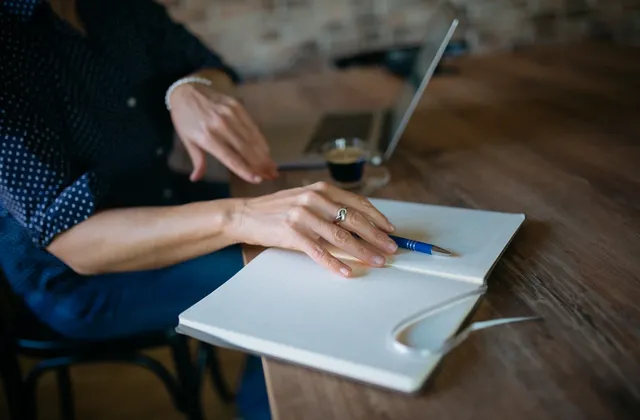 Hands resting beside a blue pen on an open notebook, with a laptop and coffee behind, photographed in November 2021