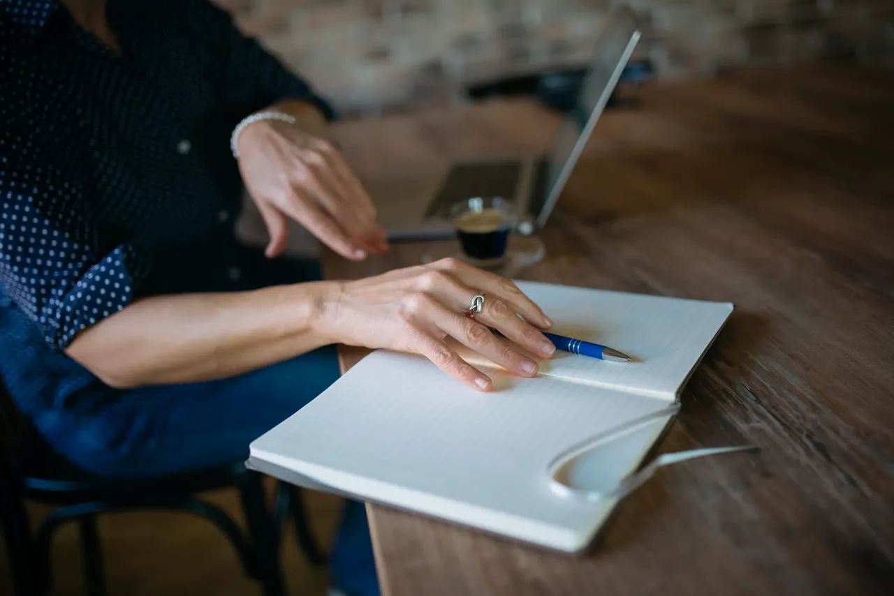 Hands resting beside a blue pen on an open notebook, with a laptop and coffee behind, photographed in November 2021