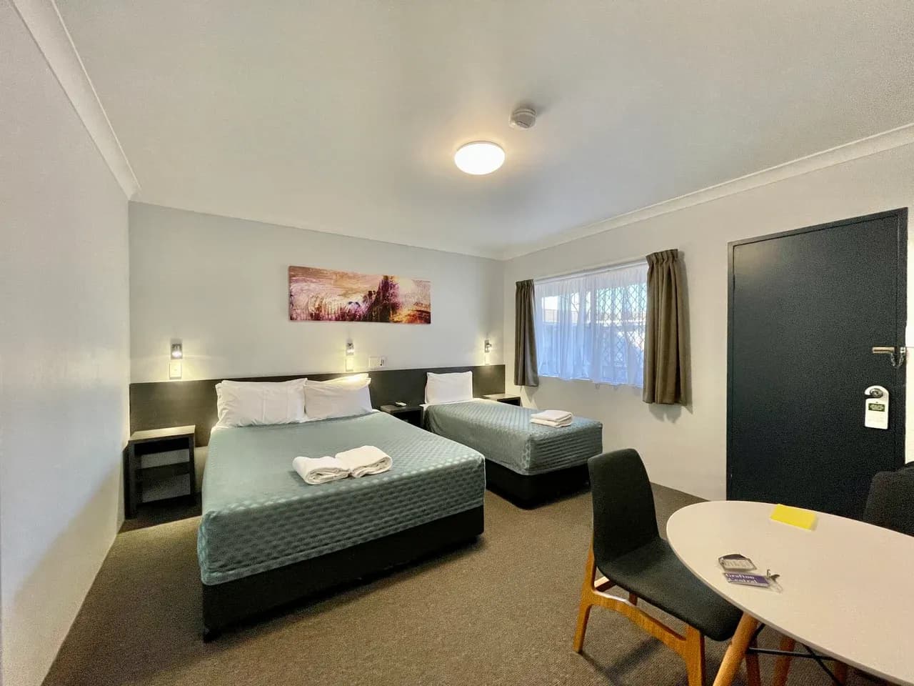 Furnished bedroom with a double bed, a single bed, a round table and a closed dark door in Grafton, New South Wales, Australia