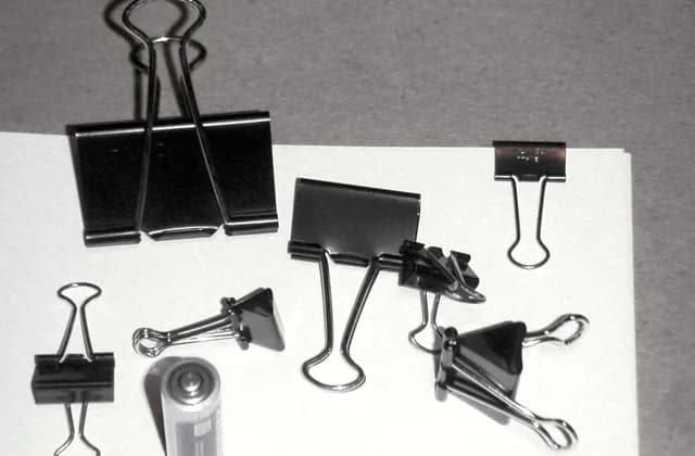 Assorted black binder clips beside a cylindrical battery on a light surface