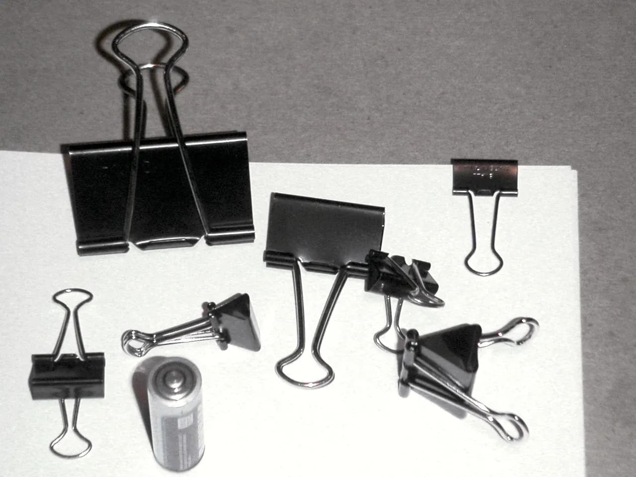 Assorted black binder clips beside a cylindrical battery on a light surface