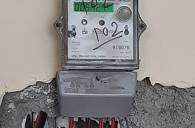 Wall-mounted electricity meter with a green display and cables below in an Indian household, photographed in July 2022