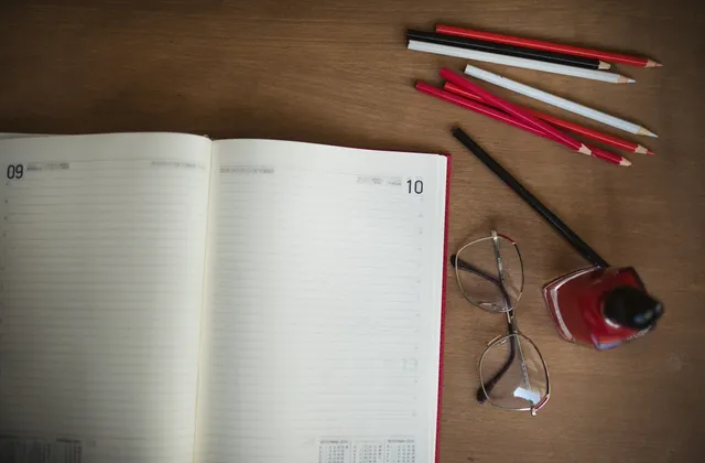 Open dated planner with pencils, glasses and an ink bottle on a wooden desk