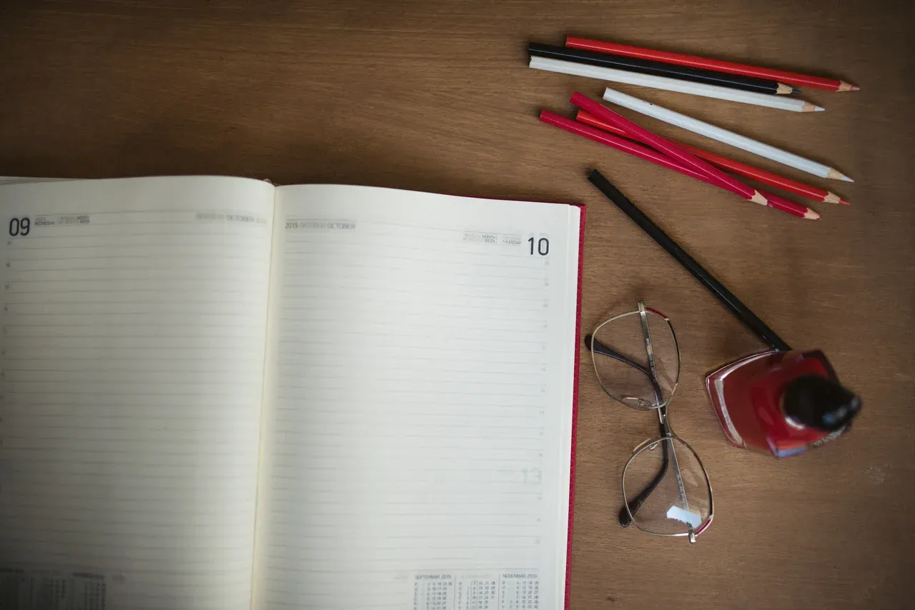 Open dated planner with pencils, glasses and an ink bottle on a wooden desk