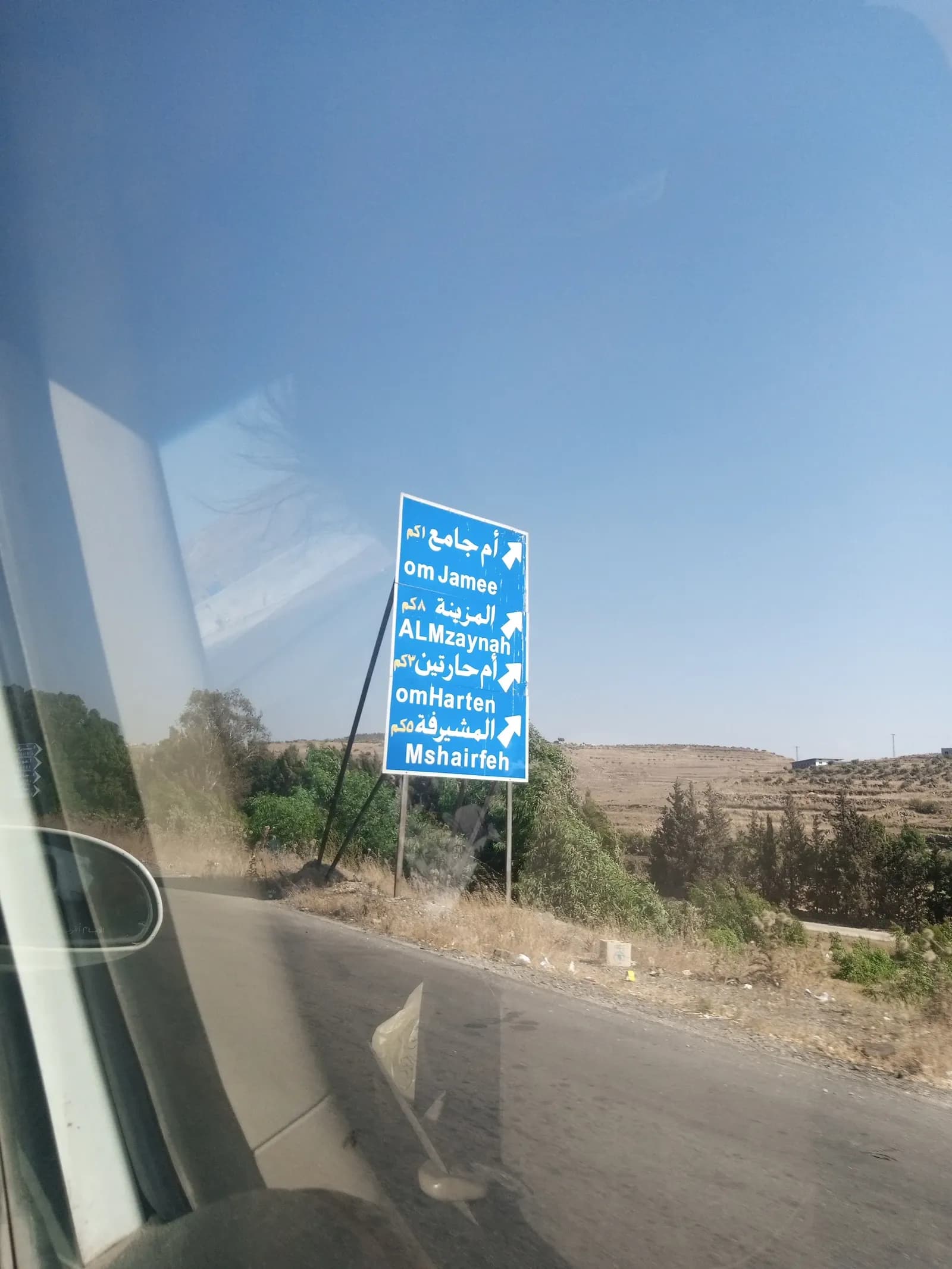Blue Arabic and English direction sign near Umm Jamee Bridge in western Homs countryside, photographed through a vehicle window in July 2024