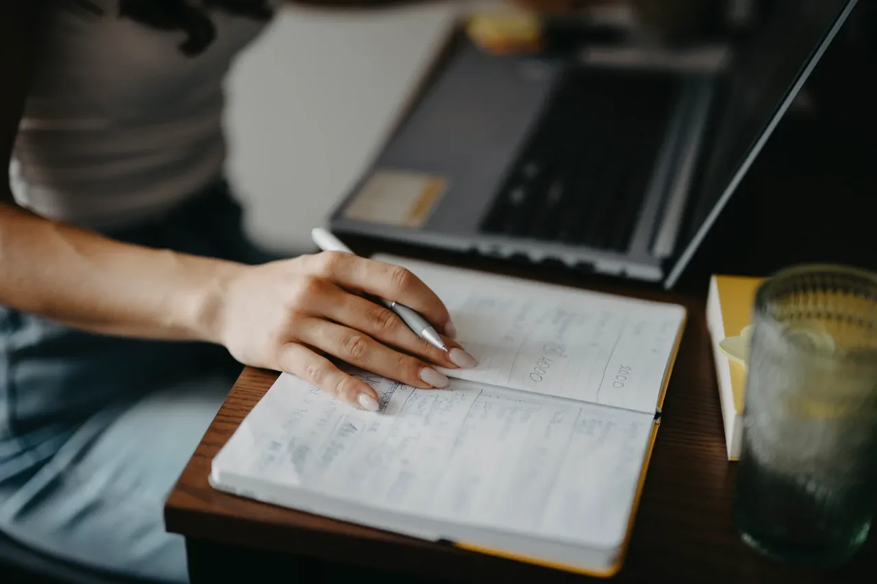 Hand writing in an open notebook beside a laptop and drinking glass, photographed in June 2026